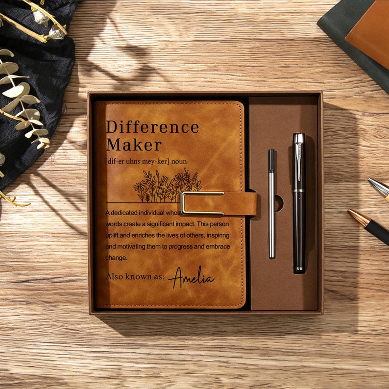 May include: A tan leather-look journal with a gold metal clasp, featuring the printed text Difference Maker, a floral line drawing, and the name Amelia. The journal is housed in a brown gift box alongside a black pen and a silver ink refill.