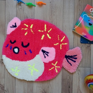 Könnte beinhalten: Ein runder, rosa fischförmiger Teppich mit weißem Bauch, rosa Flossen und gelben Sternen-Akzenten. Der Teppich hat ein cartoonartiges Design mit geschlossenen Augen. Ein Kinderbuch mit einer Bärenillustration befindet sich in der Nähe.