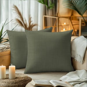 May include: Two olive green square throw pillows with a textured appearance are arranged on a beige sofa. The pillows are the focal point, with a neutral background including candles, a woven mat, and a book.
