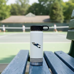 May include: White insulated water bottle with a black lid and handle, featuring a tennis player graphic in blue and black. The bottle has black stripes and a silver base. The background shows a tennis court and a wooden bench.