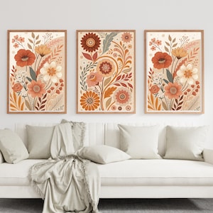 Op de afbeelding: Drie ingelijste bloemenkunstprints. Elke print toont een gestileerd botanisch ontwerp met bloemen in tinten oranje, roest en crème, tegen een beige achtergrond. De frames zijn lichtbruin van kleur. De prints worden weergegeven boven een witte bank met kussens en een deken.
