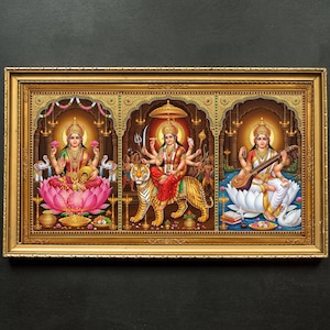 Puede incluir: Una impresión enmarcada que presenta tres deidades hindúes. El panel izquierdo muestra a Lakshmi sentada en un loto, el panel central representa a Durga montada en un tigre y el panel derecho muestra a Saraswati tocando una veena. El marco es dorado.