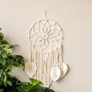 May include: Cream-colored macrame dream catcher with a circular frame and intricate web design. Features hanging tassels and leaf-shaped accents with wooden bead details. A decorative wall hanging for bohemian-style decor.