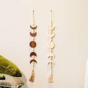 May include: Two wooden moon phase wall hangings. One is made of dark brown wood and the other is made of light wood. Both have a string of wooden beads and a tassel at the bottom. The moons are in different phases.