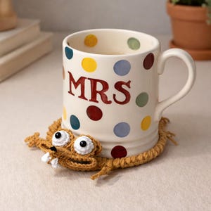 Puede incluir: Una taza de cerámica color crema con la palabra "MRS" en letras rojas y lunares de colores. La taza se apoya en un posavasos marrón tejido en forma de animal de dibujos animados con ojos y dientes blancos.
