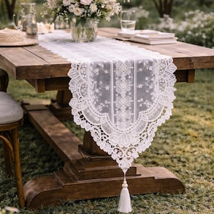 May include: A white lace table runner with a floral design and a tassel hangs from a wooden table. A bouquet of flowers sits in a glass vase. A straw hat, books, and a glass of water are also on the table.