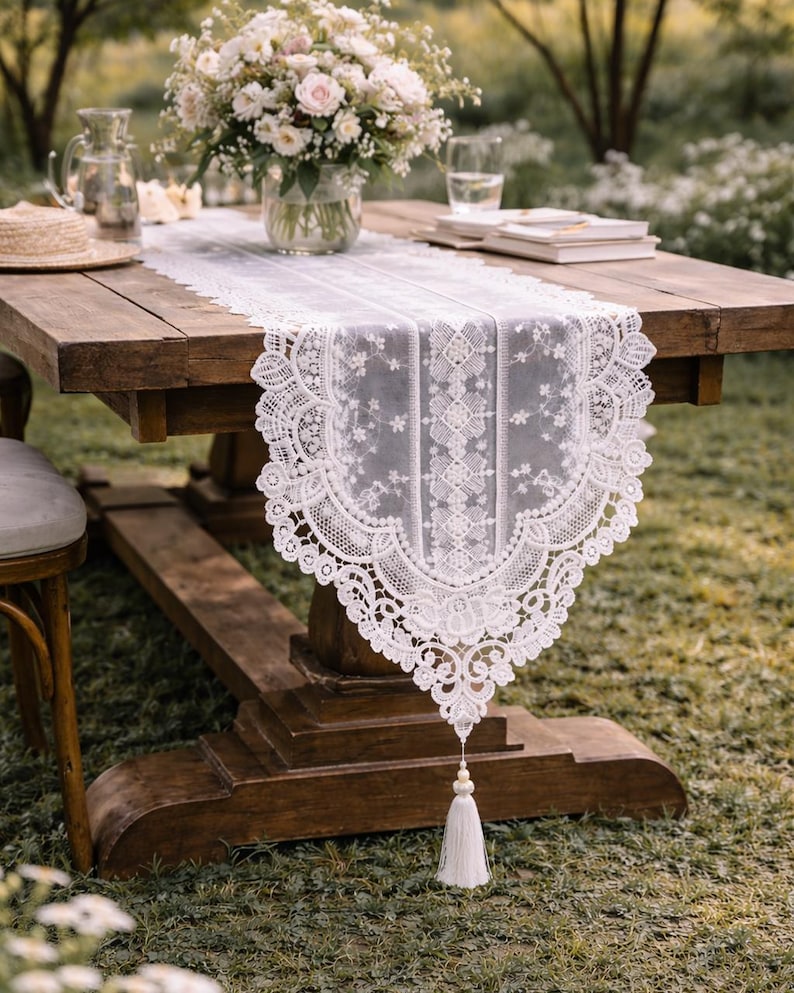 May include: A white lace table runner with a floral design and a tassel hangs from a wooden table. A bouquet of flowers sits in a glass vase. A straw hat, books, and a glass of water are also on the table.