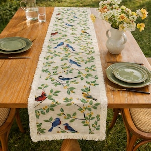 May include: A rectangular off-white table runner with a bird and floral design. The runner features various birds in shades of blue, red, and yellow perched on branches with green leaves and yellow flowers. The edges have a fringed trim. The runner is placed on a wooden table.
