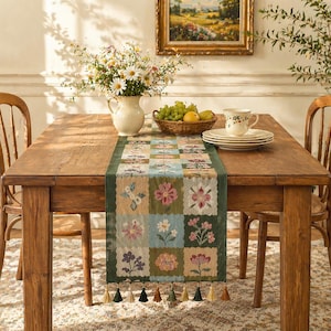 Puede incluir: Una mesa de comedor de madera está adornada con un camino de mesa con estampado floral. El camino de mesa presenta un diseño de patchwork con varios motivos florales en tonos rosa, azul y verde. Un jarrón con flores, fruta y una taza y platillo completan el conjunto.