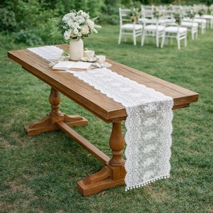 Puede incluir: Una mesa de madera con un camino de mesa de encaje blanco, un jarrón con flores blancas y dos tazas de té. La mesa está instalada al aire libre sobre una zona de césped, con sillas blancas al fondo. La mesa mide unos 152 cm de largo.