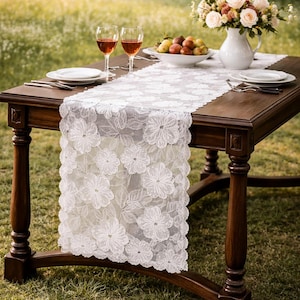 May include: A dark wood dining table is set for a meal with a white floral lace table runner. Two wine glasses with red wine, white plates, silverware, a bowl of fruit, and a vase of flowers complete the setting. The table is outdoors.