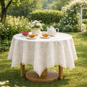 May include: A round wooden table set for tea outdoors. The table is covered with a white, floral-patterned tablecloth. A white teapot, two teacups, a vase of flowers, and a bowl of berries are arranged on the table.