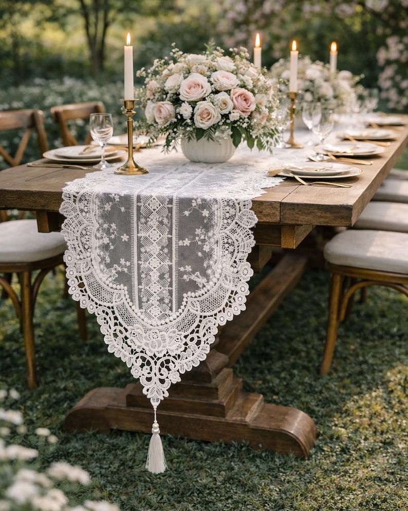 May include: A wooden dining table set for a special occasion, featuring a white lace table runner with a tassel, floral centerpieces, candles, and place settings. The table is surrounded by wooden chairs, set outdoors on a grassy area.