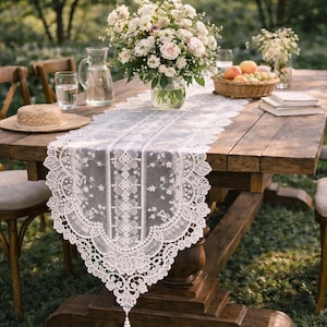 May include: A white lace table runner with a floral design is displayed on a rustic wooden table. The table is set for an outdoor gathering, with flowers, fruit, and a straw hat. The runner has a scalloped edge.