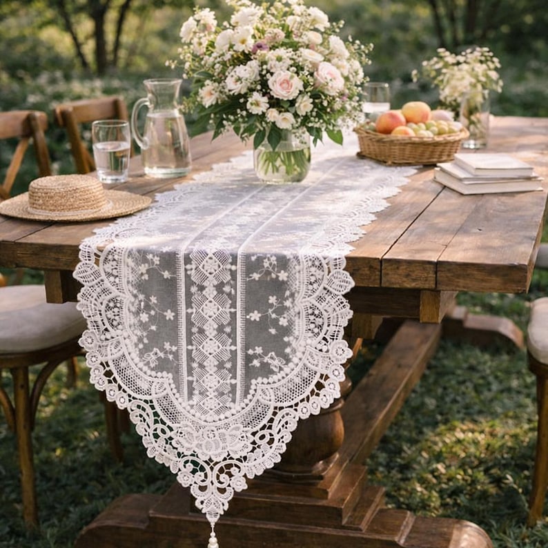 May include: A white lace table runner with a floral design is displayed on a rustic wooden table. The table is set for an outdoor gathering, with flowers, fruit, and a straw hat. The runner has a scalloped edge.