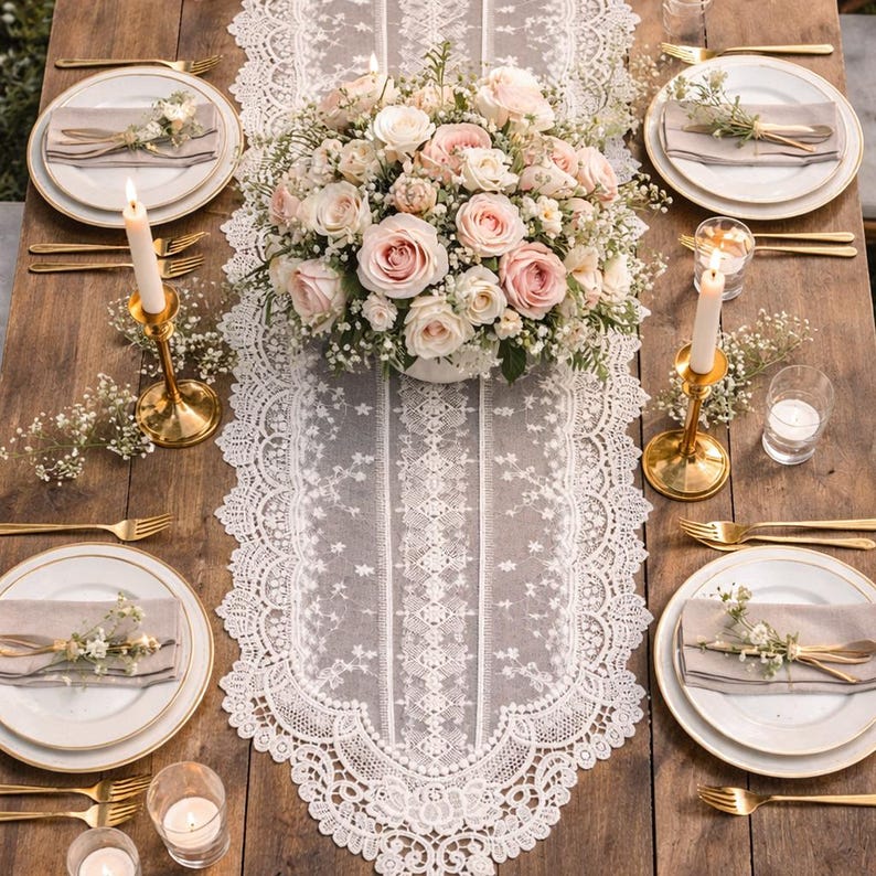 May include: A beautifully set table with a white lace runner, gold-rimmed plates, and gold cutlery. A centerpiece of pink and white roses sits atop the runner. Gold candle holders with lit candles and small floral arrangements complete the elegant setting.