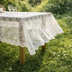 Peut inclure: Une nappe en dentelle blanche à motifs floraux recouvre une table en bois. La nappe a un bord festonné et retombe sur les côtés de la table. La table est dressée à l'extérieur sur une pelouse.