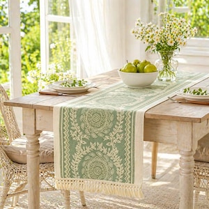 May include: A green and cream table runner with a floral design and fringed edges. It's laid on a wooden table set for a meal, with plates, cutlery, and a bowl of green pears. A vase of white flowers is also on the table.