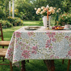 Op de afbeelding: Een rechthoekig tafelkleed met bloemenpatroon, met roze bloemen en groene accenten, bedekt een houten tafel. Een witte vaas met perzikrozen, kaarsen en borden staan op de tafel. De scène speelt zich buiten af in een tuin.