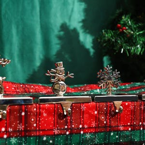 May include: Four silver Christmas stocking holders featuring a reindeer, snowman, snowflake, and Christmas tree. They are displayed on a red and green plaid surface, with a Christmas tree visible in the background.