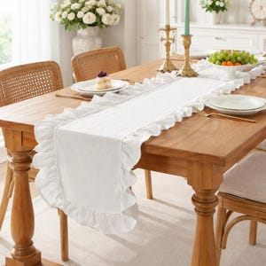 May include: A wooden dining table is set with a white ruffled table runner. The table is adorned with a white floral arrangement, candles, and a bowl of fruit. Wicker chairs surround the table, creating a warm and inviting atmosphere.