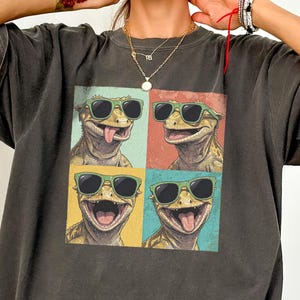 May include: A charcoal grey short-sleeve t-shirt featuring a graphic print of four crested geckos wearing green sunglasses. The geckos are arranged in a grid pattern with individual backgrounds in light blue, red, yellow, and teal colours.