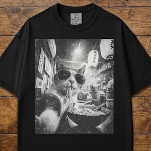 May include: Black t-shirt featuring a grayscale image of a cat wearing sunglasses and eating ramen noodles. The cat appears to be taking a selfie in a Japanese restaurant setting. The shirt has a Comfort Colors tag.