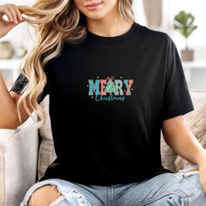 K&ouml;nnte beinhalten: Schwarzes T-Shirt mit einem farbenfrohen "Merry Christmas"-Motiv. Das Design zeigt einen Weihnachtsbaum und Text in T&uuml;rkis, Rosa und Blau. Das Shirt ist aus weichem Material und hat kurze &Auml;rmel.