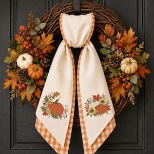 May include: A grapevine wreath decorated with orange and yellow maple leaves, pinecones, berries, and small white and orange pumpkins. A cream-colored fabric sash with orange gingham trim hangs from the center, featuring embroidered pumpkin designs.