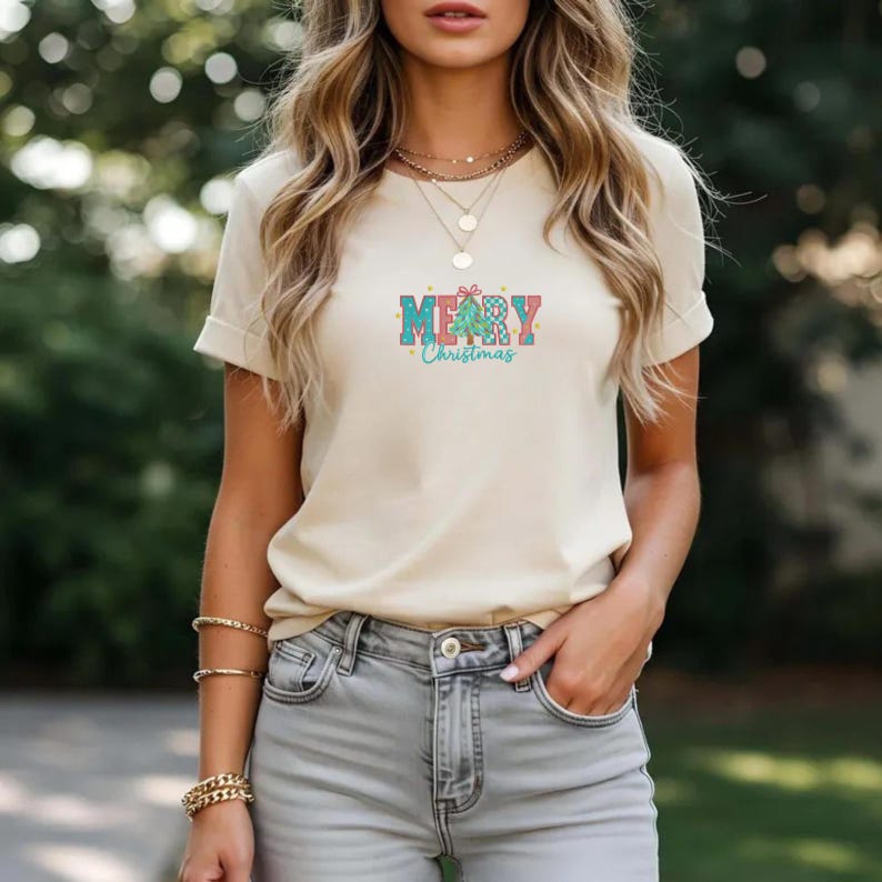 K&ouml;nnte beinhalten: Cremefarbenes T-Shirt mit der Aufschrift "MERRY Christmas" in einem farbenfrohen, festlichen Design. Der Text enth&auml;lt eine Weihnachtsbaum-Grafik. Das Shirt ist kurz&auml;rmlig und hat einen Rundhalsausschnitt.