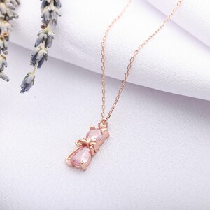 May include: A rose gold cat pendant necklace with pink gemstone accents. The cat pendant features a bow and two pink gemstones. The necklace is displayed on a white background with lavender sprigs.
