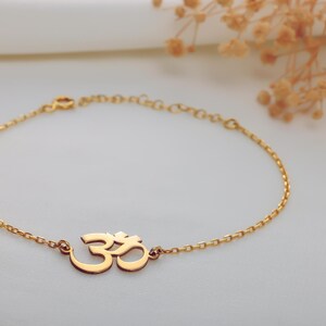 Pode incluir: Pulseira dourada com o símbolo Om. A delicada corrente está conectada ao símbolo Om, que é o ponto focal da pulseira. A pulseira é exibida em uma superfície branca, com um fundo macio e desfocado.