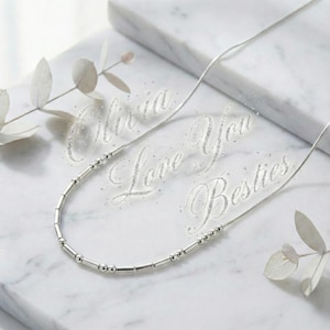 May include: A silver necklace featuring a series of small, round beads and cylindrical bars. The necklace is presented on a white marble surface, with the words "Olivia Love You Besties" in a glittery script.