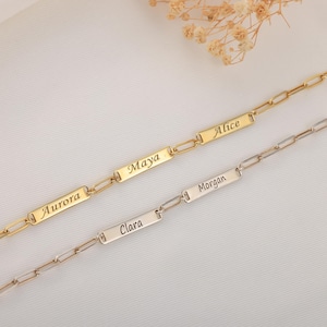 Può includere: Due braccialetti a catena a maglie personalizzati. Uno è dorato, l'altro argentato. Ogni braccialetto presenta targhette rettangolari con nomi incisi: Aurora, Maya, Alice, Clara e Morgan. I braccialetti sono esposti su una superficie bianca.