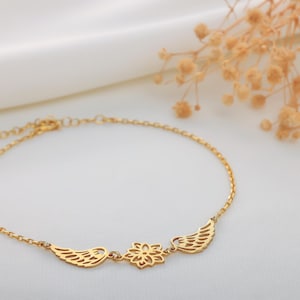 May include: A delicate gold-colored bracelet featuring a floral design in the center, flanked by two wing-shaped charms. The bracelet is displayed on a white surface, with dried flowers in the background.