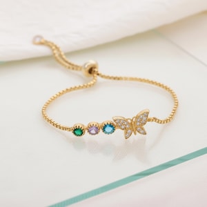 May include: A gold-colored bracelet featuring a butterfly and three colored gemstones. The butterfly is adorned with small, clear crystals. The bracelet has an adjustable sliding clasp. The gemstones are green, purple, and blue.