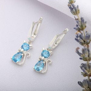 May include: A pair of silver cat-shaped earrings with blue gemstone accents. Each earring features a round gemstone head and a teardrop-shaped gemstone body, with a curved tail design. The earrings are set against a white background.