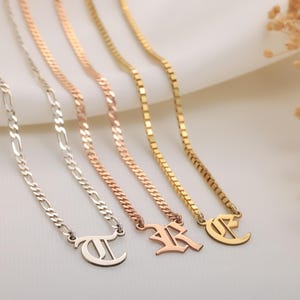 May include: Four necklaces with initial pendants in silver, rose gold, and gold. The chains have different styles. The pendants are shaped like the letters T, R, and C. The necklaces are displayed on a white background.