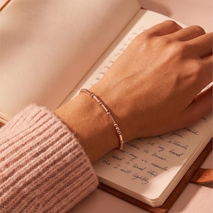 Op de afbeelding: Een roségouden armband met een reeks kleine, ronde kralen en langwerpige, staafvormige schakels. De armband wordt om een pols gedragen en rust op een open notitieboek met handgeschreven tekst. De persoon draagt een roze trui.