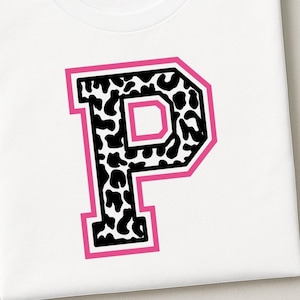 May include: A white t-shirt featuring a large, block-style letter P centered on the chest. The letter has a black and white cow print pattern fill, outlined with a thick, vibrant pink border that gives it a bold, collegiate-inspired appearance.