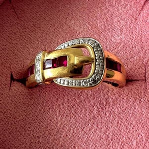 May include: A gold-coloured ring shaped like a belt buckle, featuring rectangular ruby gemstones and diamond detailing. The ring is set against a pink background.