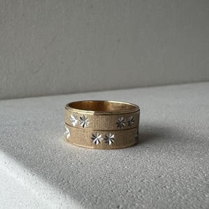 May include: A gold ring with a textured finish, featuring a band of star-shaped cutouts. The ring is made of metal and rests on a white surface. This is a piece of jewellery.
