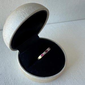 Puede incluir: Un anillo de banda de oro con piedras rojas y claras alternas, presentado en un joyero abierto de color crema con interior negro. El anillo es el punto focal, destacando su diseño.