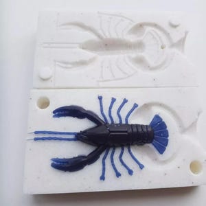 May include: A black and blue plastic crawfish lure sits on top of a white mold. The crawfish has blue claws, legs, and tail. The mold is rectangular with a crawfish impression.