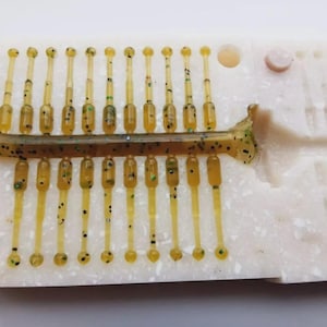 May include: A close-up of a white mould holding several small, clear, yellow fishing lures. The lures have a thin design with a rounded end and are speckled with black and green dots. A larger, similar lure is also visible.