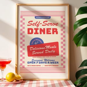 May include: A framed poster with a cream background, pink checkered borders, and red text reading Self-Serve DINER, Delicious Meals Served Daily, and OPEN 7 DAYS A WEEK. The poster includes blue accents and text, with a red cocktail and citrus fruit nearby.