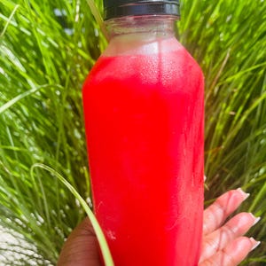 May include: A clear plastic bottle filled with a vibrant red liquid, topped with a black cap. The bottle is held in a hand against a backdrop of green foliage, suggesting a refreshing beverage.