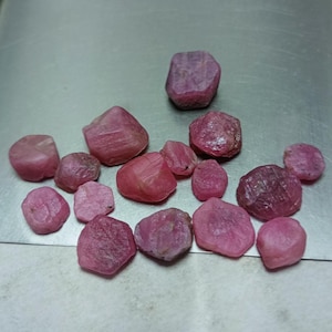 Peut inclure: Une collection de pierres précieuses de rubis brutes, non taillées, de tailles et de formes variées. Les rubis présentent une gamme de teintes allant du rose au rouge profond, avec une surface rugueuse et texturée. Ces pierres naturelles sont disposées sur une surface métallique.