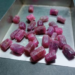 May include: A collection of raw, uncut ruby gemstones in varying sizes and shapes. The gemstones are a deep pinkish-red color and have a rough, textured surface. They are displayed in a silver metal tray.