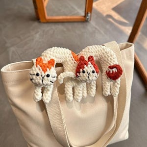 Könnte beinhalten: Eine cremefarbene Tragetasche mit zwei gehäkelten Katzenverzierungen. Die Katzen sind weiß mit orangefarbenen und roten Akzenten. Die Tasche hat ein schlichtes Design und einen Schulterriemen.